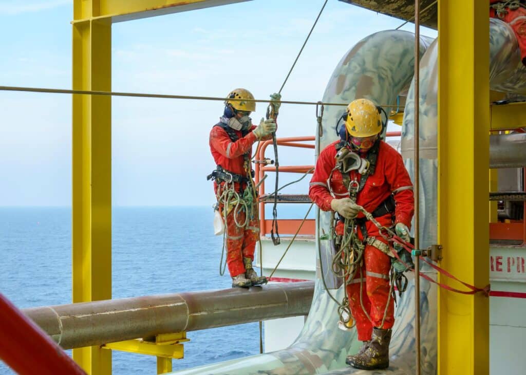 Working at height. A group of abseilers wearing red coverall and personal protective equipment (ppe) standing on the piepeline arranging their rope access with background open sea.