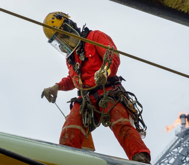 Working at height. An abseiler wearing personal protective equipment (ppe) standing on pipeline for painting with background flare tip burning in the sky.