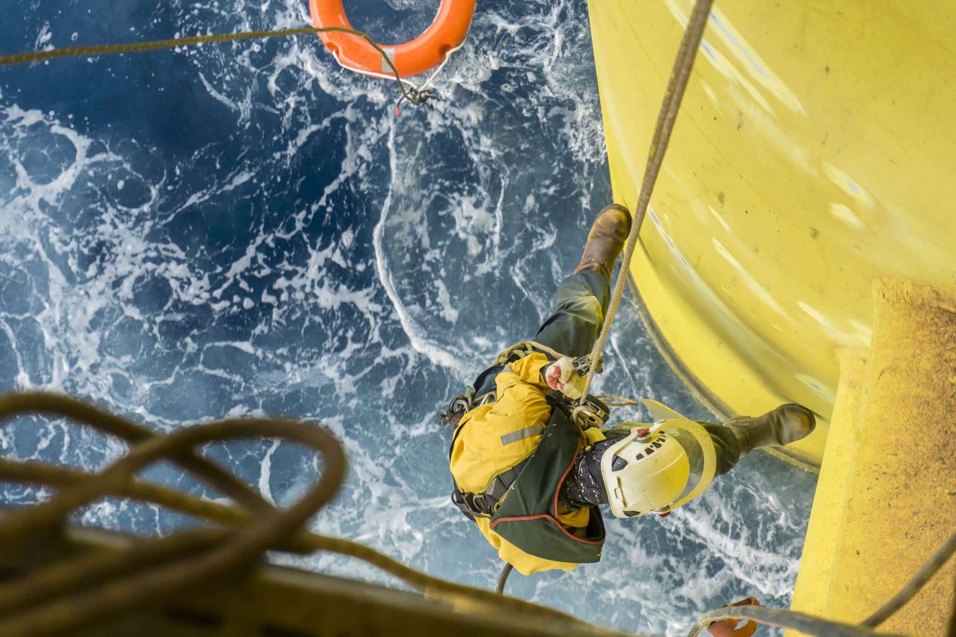 Working overboard. A commercial abseiler complete with personal protective equipment (ppe) hanging at oil and gas platform jacket module while life buoy standby on sea surface.
