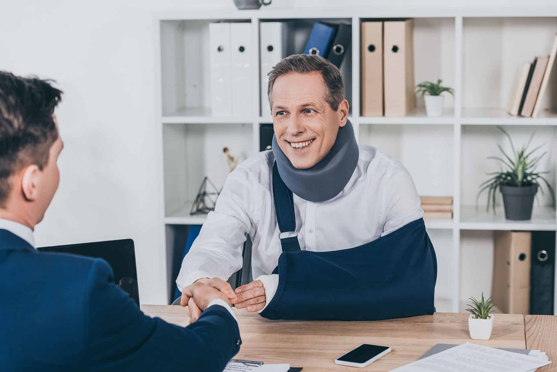 Worker in neck brace with brokenarm and businessman in blue jacket shaking hands over table in office