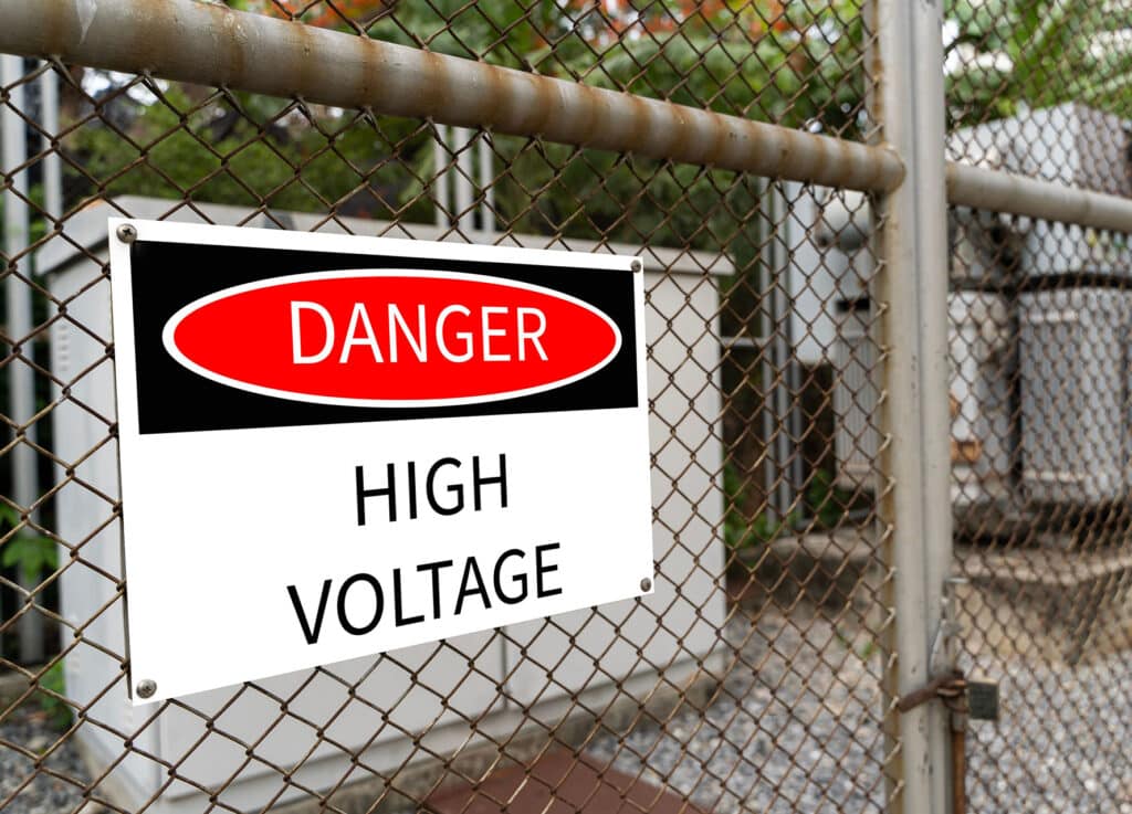 High-voltage transformer substation behind barbed-wire chain-link fence with danger high voltage sign.