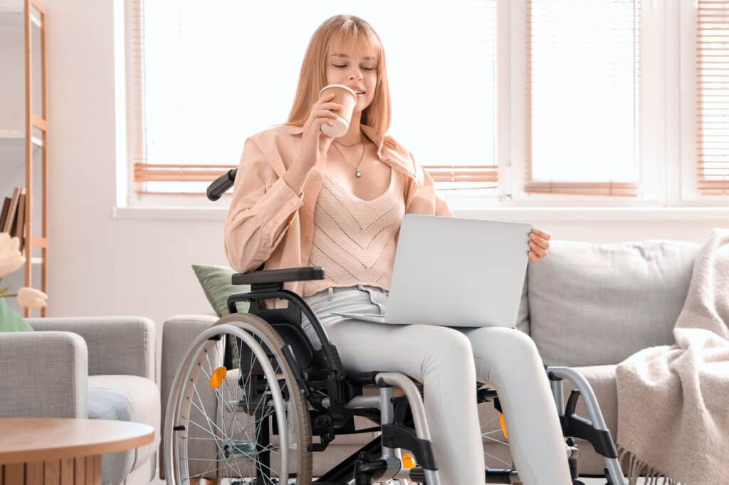 Young woman in wheelchair with laptop drinking coffee at home