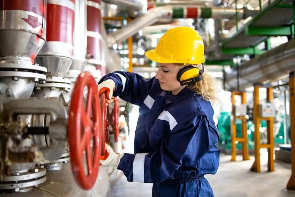 Professional refinery worker in protective uniform and hardhat standing by natural gas pipes and closing valve.