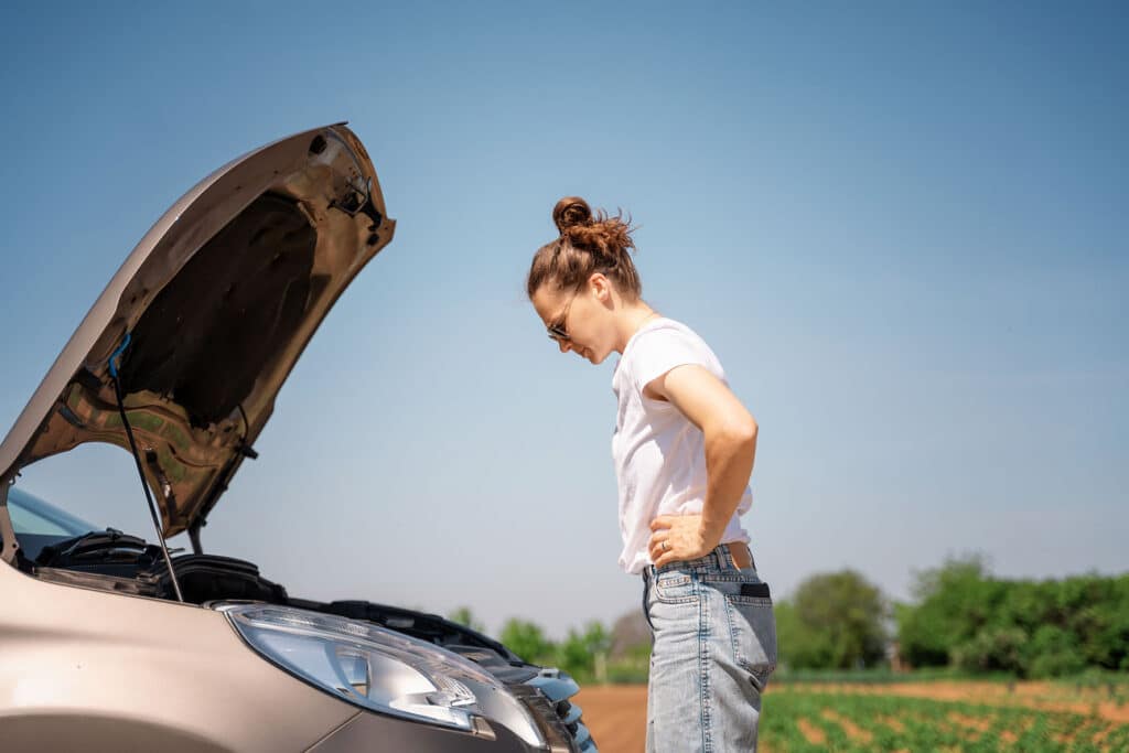 Young woman driver standing by the open hood of a car in the countryside