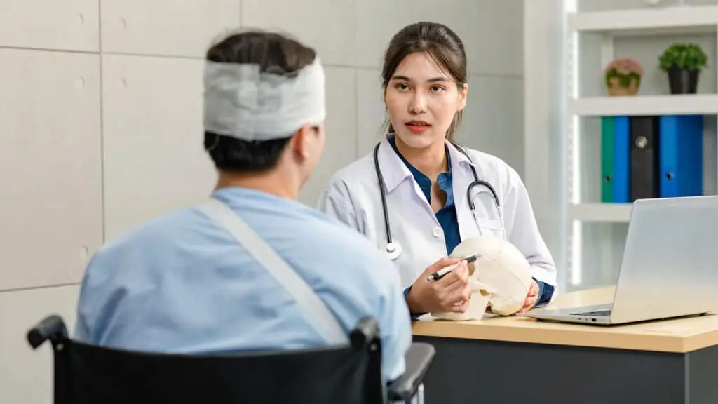 Asian young professional female practitioner doctor in white lab coat with stethoscope holding skull model showing explaining to unrecognizable male patient sitting on wheelchair after brain surgery.