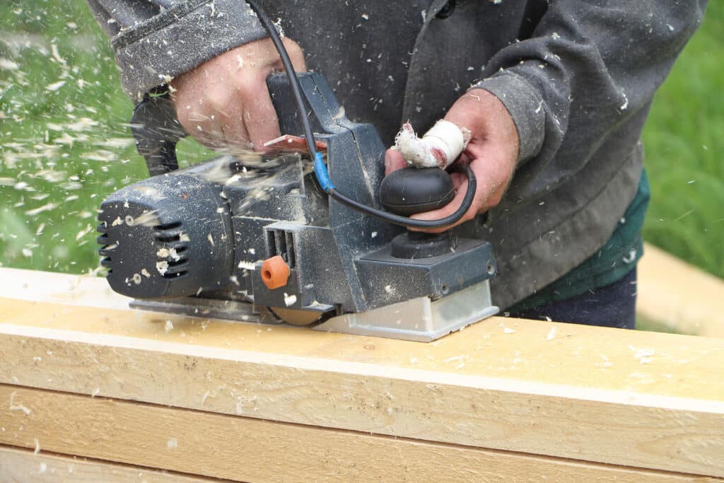 The man with the injured finger processes a board a jointer plane outdoors