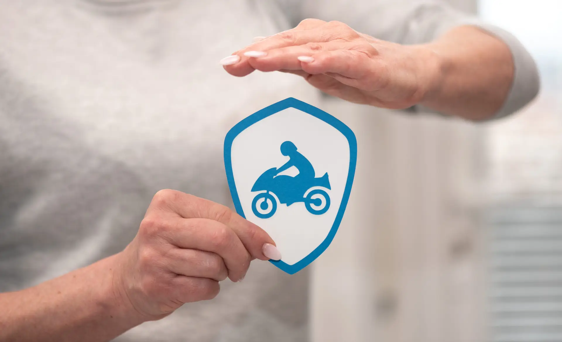 a motorcycle, blue, on a white shield with hands covering them symbolizing motorcycle insurance