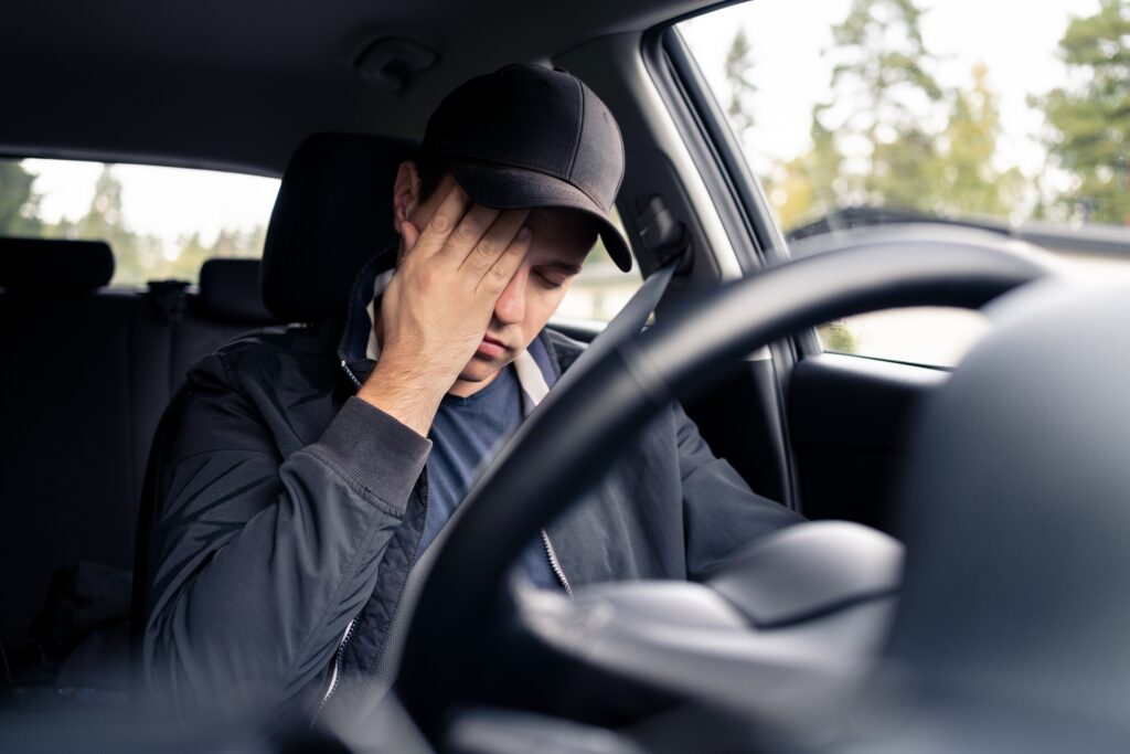 a man sitting behind the steering wheel of a vehicle rubbing his face because he is drowsy and falling asleep