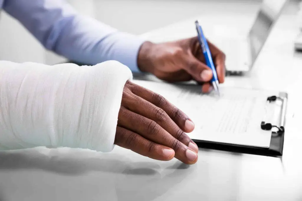 Overhead view of injured man with bandage hand filling insurance claim form on clipboard