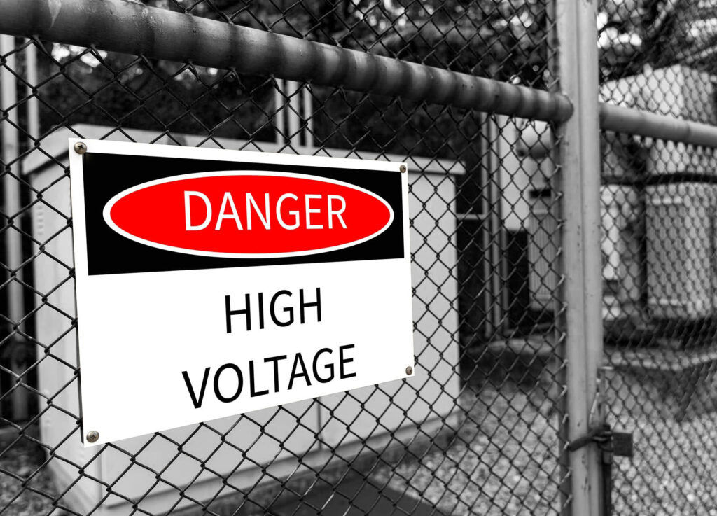 High-voltage transformer substation behind barbed-wire chain-link fence with danger high voltage sign.